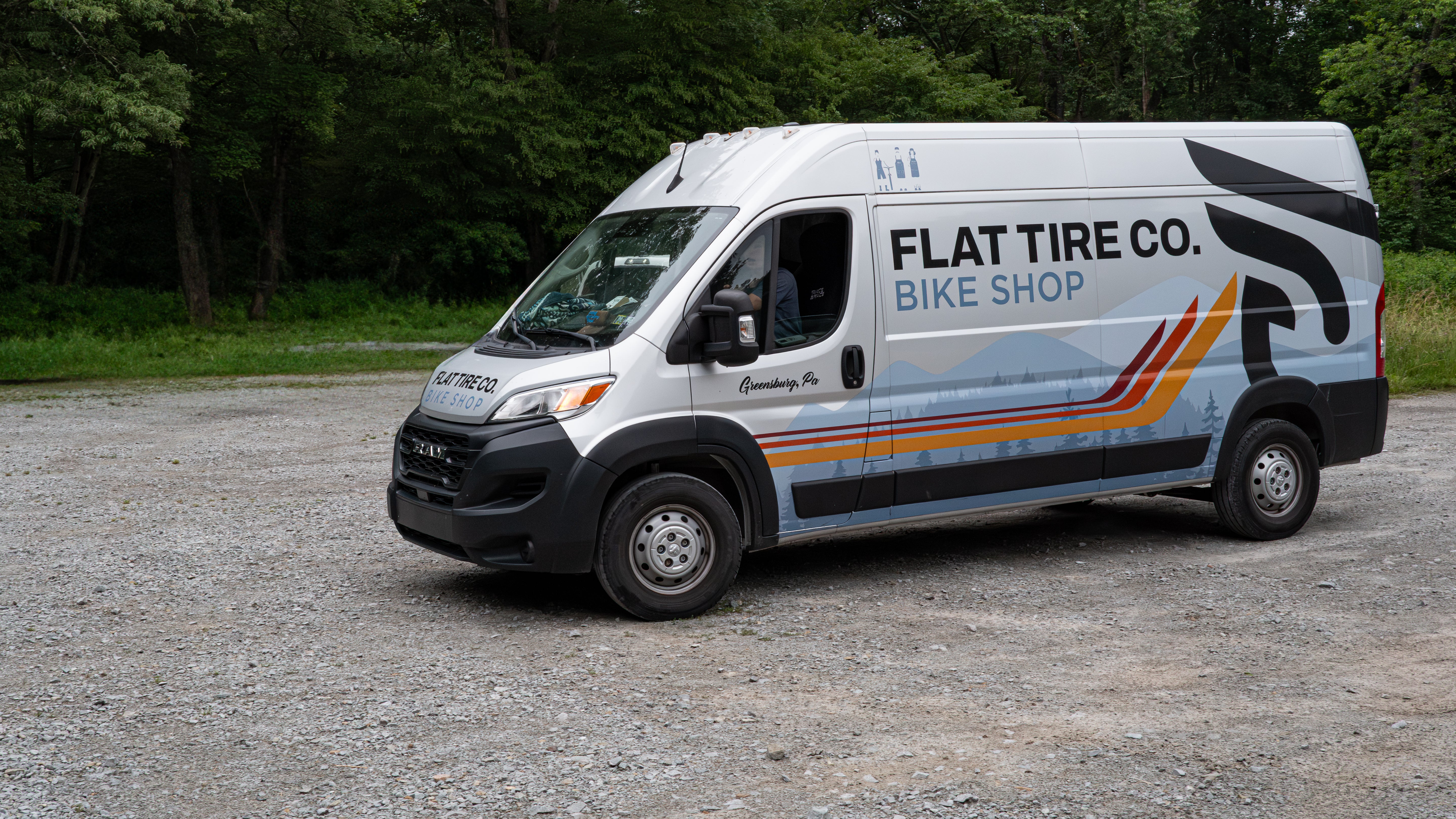 Van with 'Flat Tire Co. Bike Shop' branding parked on a gravel road.